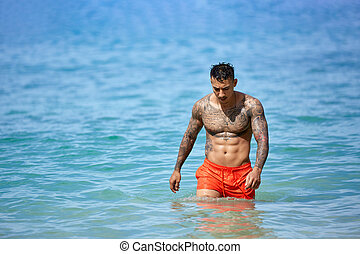 athletic-tattooed-guy-walking-through-the-water-at-the-tropical-beach-athletic-shirtless-tattooed-picture_csp89259218
